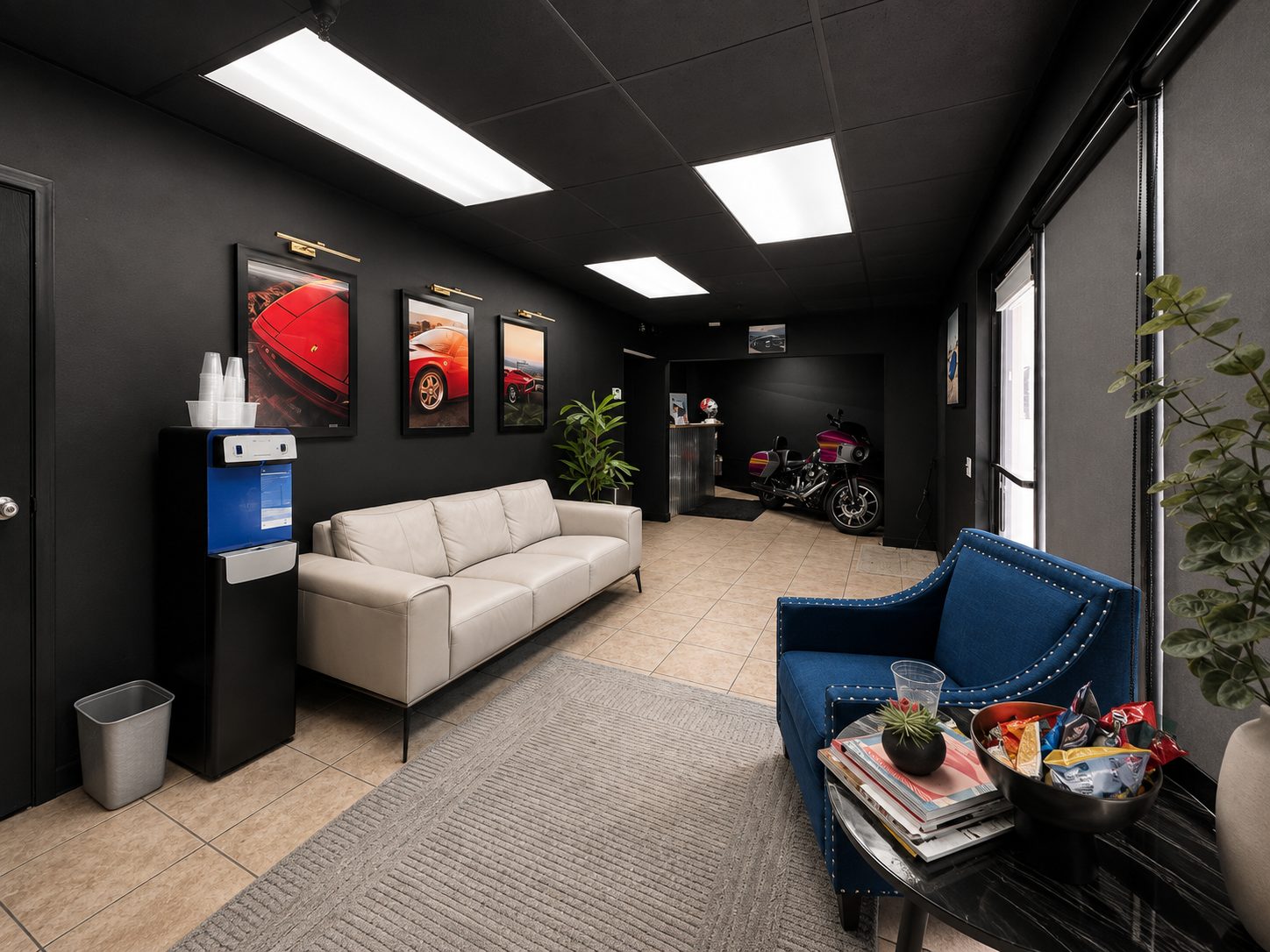 The client lounge at Gravity Garage, with a leather sofa, upholstered armchair, framed automotive prints, refreshments, and a water station.
