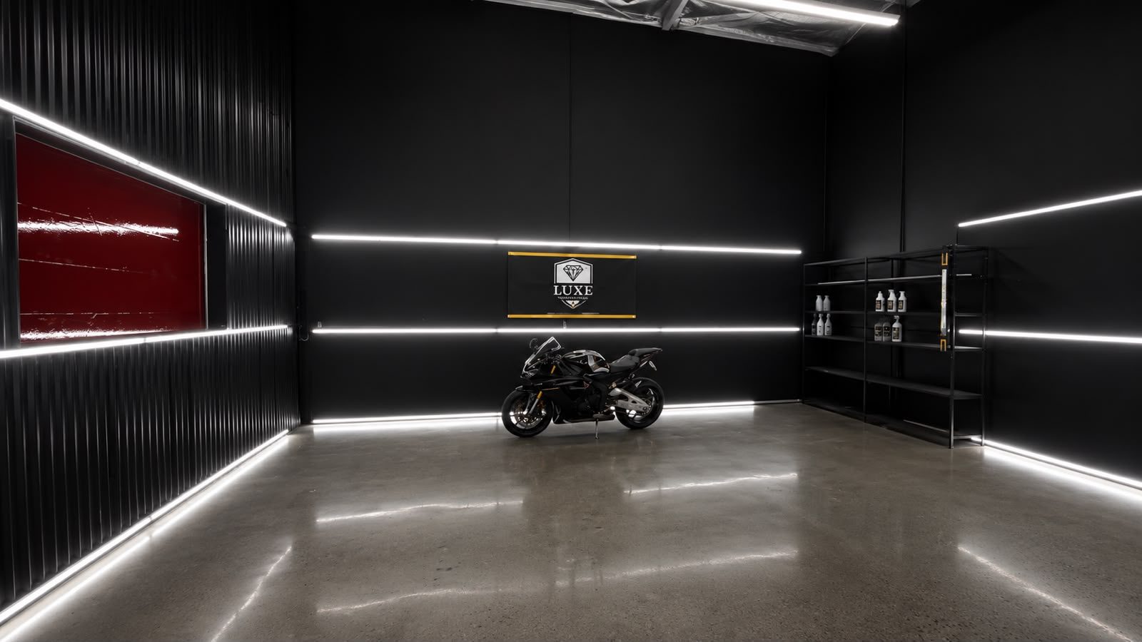 A blacked-out presentation bay at Gravity Garage lined with continuous LED strip lighting, a motorcycle parked in the center on polished concrete, a LUXE Protection Films banner on the far wall and shelving stocked with product to one side.