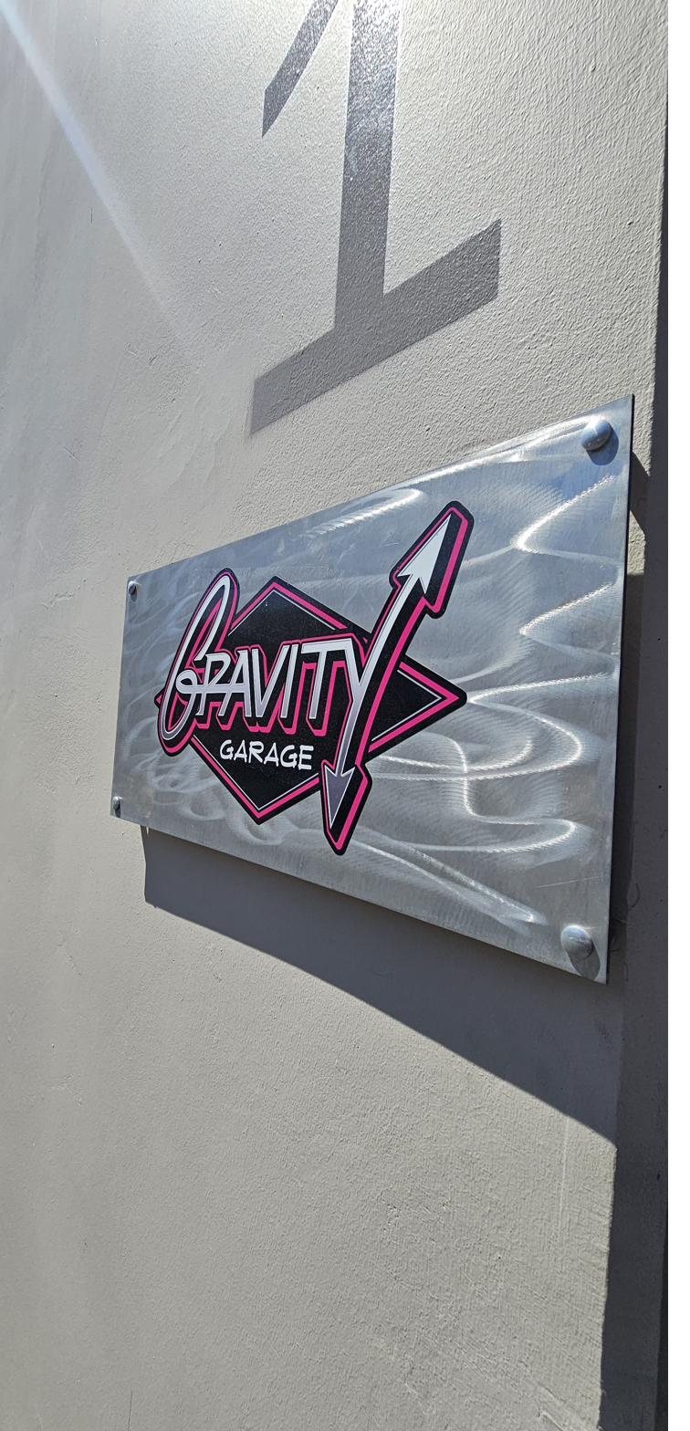 The Gravity Garage logo on a brushed metal sign plate mounted beside the unit entrance, lit by direct sunlight.