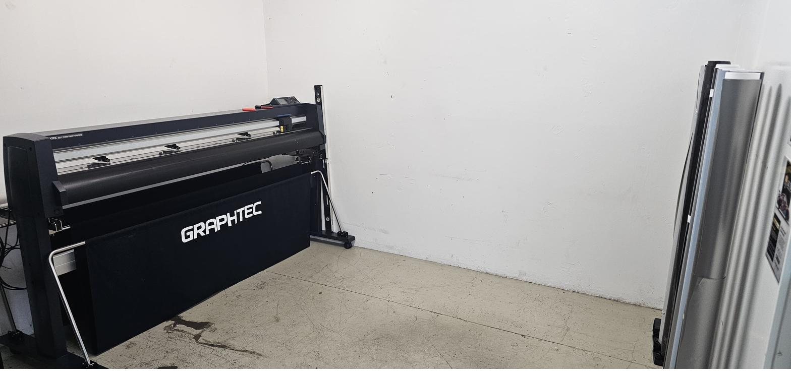 A Graphtec Cutting Pro FC9000 wide-format cutting plotter set up inside Gravity Garage, with a vertical rack of film sample rolls standing beside it.