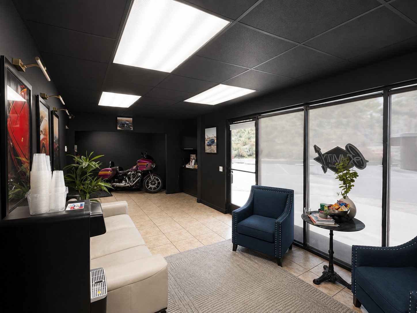 The Gravity Garage front showroom: a black-walled reception area with framed automotive artwork, a customer seating area, and a custom motorcycle displayed in an alcove beyond the reception desk. The Gravity Garage logo is applied to the front window.