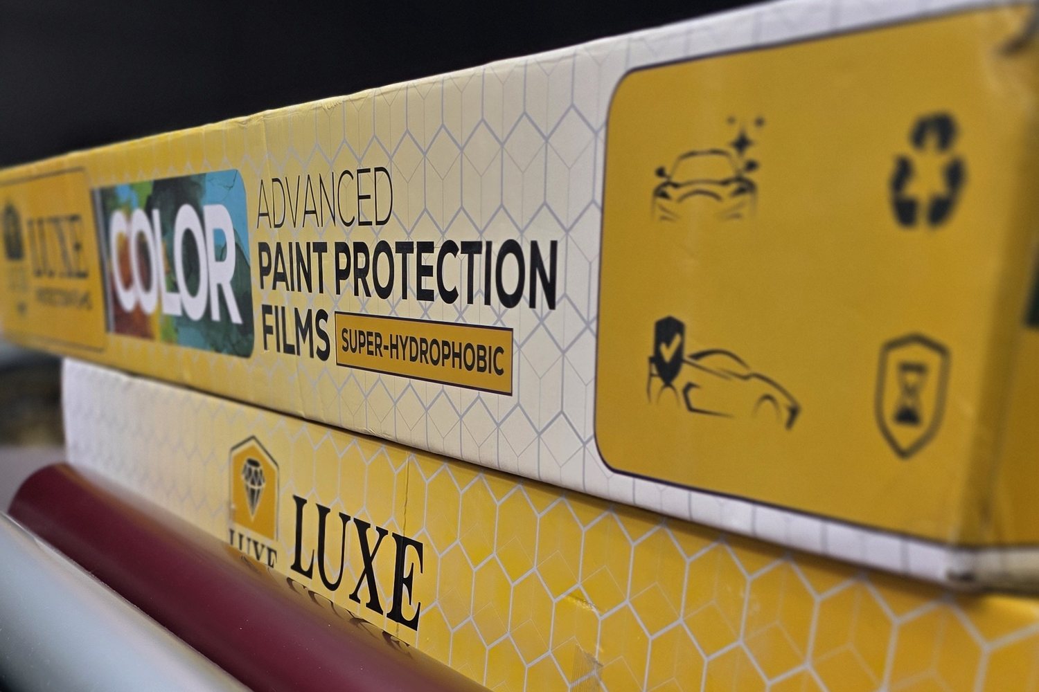 A yellow LUXE Color Series carton — Advanced Paint Protection Films, Super-Hydrophobic — stacked on a LUXE carton on the rack, with a roll of deep red film alongside.