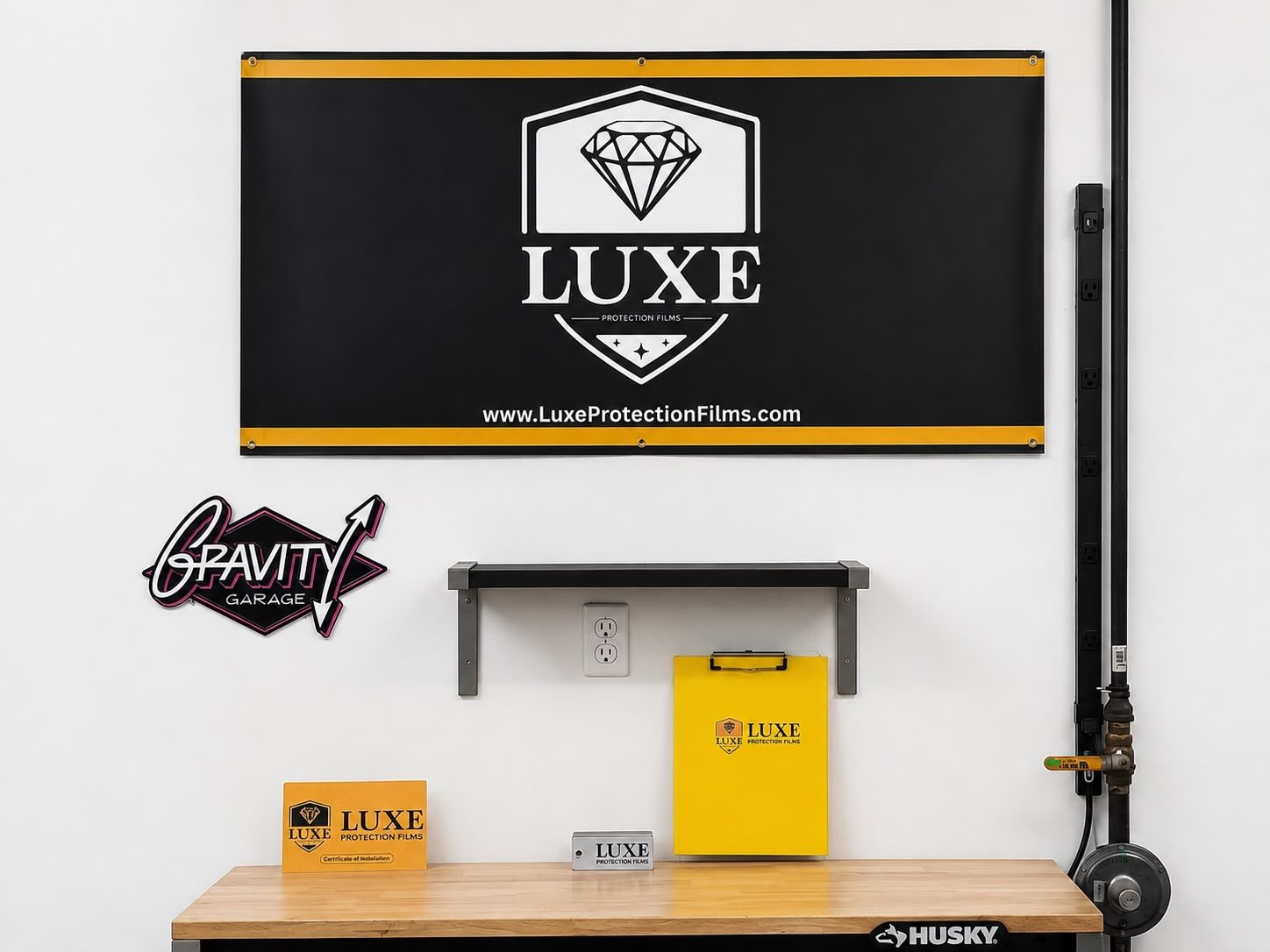 Inside Gravity Garage: a LUXE Protection Films banner mounted above a workbench, the Gravity Garage sign on the wall beside it, and a LUXE certificate of installation, branded clipboard and plaque on the bench below.