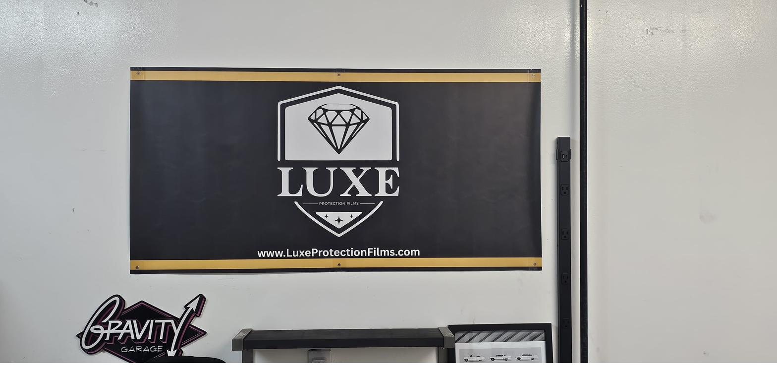 A LUXE Protection Films banner mounted on the wall inside Gravity Garage, with the Gravity Garage logo sign displayed on the same wall directly beneath it.