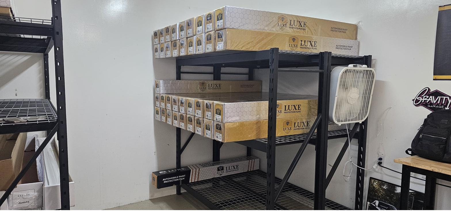 Rolls of LUXE Protection Films paint protection film in branded boxes stacked across heavy black industrial racking inside the Gravity Garage stocking area.
